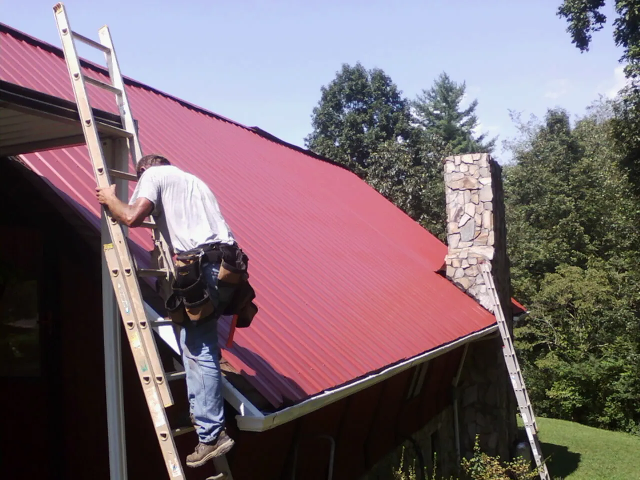 Roofer on a ladder beside a red metal roof and stone chimney — Soffit & Fascia Repair by Roofing Brothers in North Newton